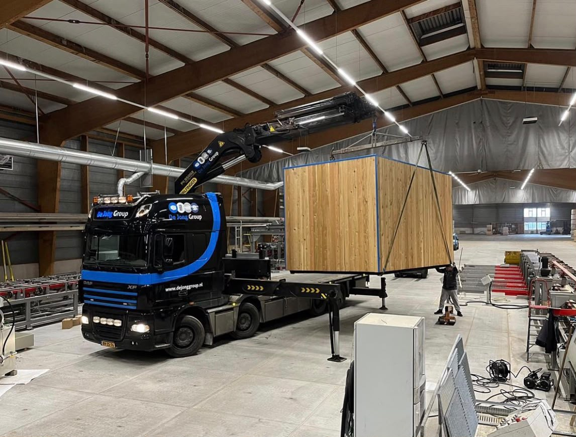 Vrachtwagen met laadkraan die een houten unit in een hal verplaatst